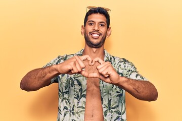 Young latin man wearing summer shirt and sunglasses smiling in love doing heart symbol shape with hands. romantic concept.