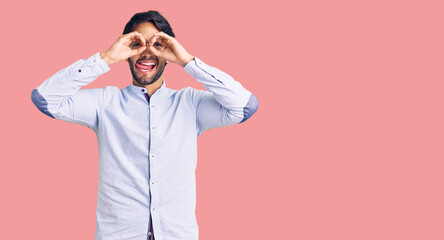Handsome hispanic man wearing business shirt and glasses doing ok gesture like binoculars sticking tongue out, eyes looking through fingers. crazy expression.