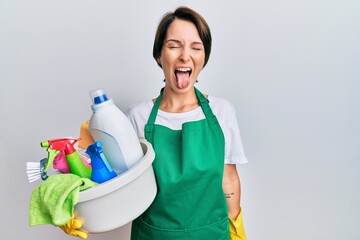 Young brunette woman with short hair wearing apron holding cleaning products sticking tongue out happy with funny expression. emotion concept.
