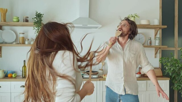 Happy Couple Dancing in Kitchen and Singing Karaoke Using Kitchenware Like Microphones While Cooking. Lovely Couple Enjoys Food Preparation Together During Romantic Date at Home.