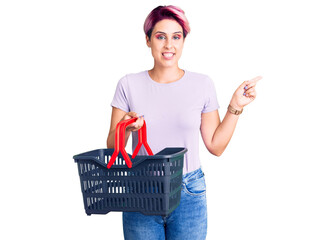 Young beautiful woman with pink hair holding supermarket shopping basket smiling happy pointing with hand and finger to the side