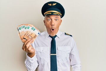 Handsome middle age mature man wearing airplane pilot uniform holding euros scared and amazed with open mouth for surprise, disbelief face