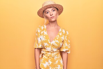 Beautiful blonde woman on vacation wearing summer hat and dress over yellow background Relaxed with serious expression on face. Simple and natural looking at the camera.