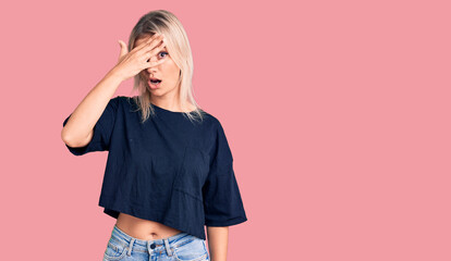 Young beautiful blonde woman wearing casual t-shirt peeking in shock covering face and eyes with hand, looking through fingers with embarrassed expression.