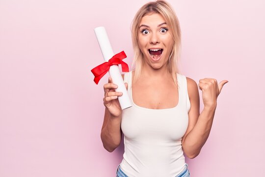 Young beautiful blonde student woman holding graduated degree diploma over pink background pointing thumb up to the side smiling happy with open mouth