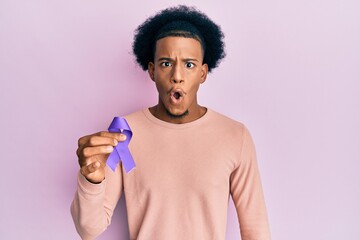 African american man with afro hair holding awareness purple ribbon scared and amazed with open...