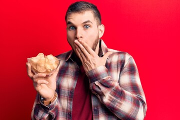 Young handsome man holding bowl with potato chips covering mouth with hand, shocked and afraid for...