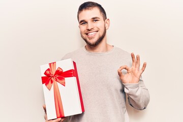 Young handsome man holding gift doing ok sign with fingers, smiling friendly gesturing excellent symbol