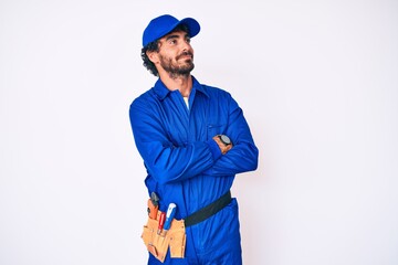 Handsome young man with curly hair and bear weaing handyman uniform looking to the side with arms crossed convinced and confident