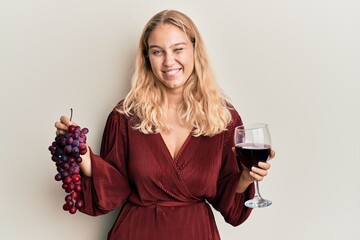 Young blonde girl holding branch of fresh grapes and red wine winking looking at the camera with...