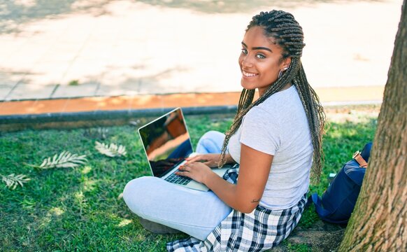Young african american student woman smiling happy using computer laptop sitting on the grass at the university campus