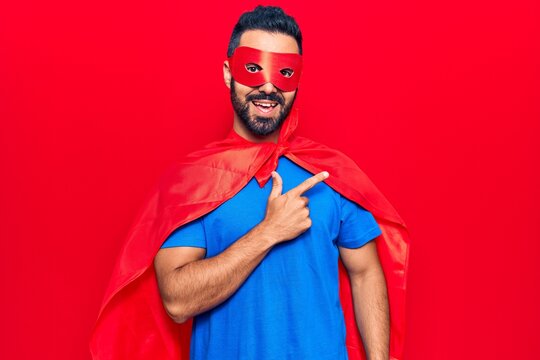 Young hispanic man wearing super hero costume smiling cheerful pointing with hand and finger up to the side