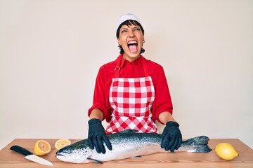 Beautiful brunettte woman fishmonger cooking fresh raw salmon angry and mad screaming frustrated and furious, shouting with anger looking up.