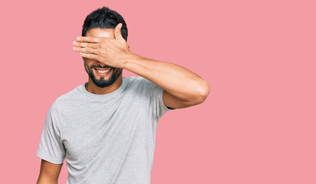 Young man with beard wearing casual grey tshirt smiling and laughing with hand on face covering eyes for surprise. blind concept.