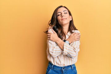 Young brunette woman wearing casual clothes over yellow background hugging oneself happy and positive, smiling confident. self love and self care