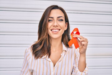 Young hispanic woman smiling happy holding awareness orange ribbon standing at the city.