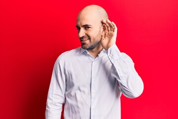 Young handsome bald man wearing elegant shirt smiling with hand over ear listening and hearing to rumor or gossip. deafness concept.