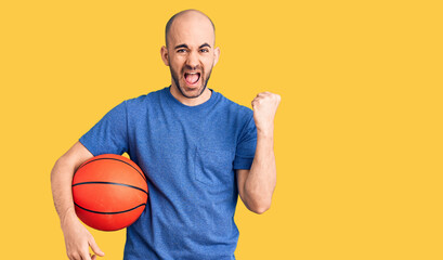 Young handsome man holding basketball ball screaming proud, celebrating victory and success very excited with raised arms