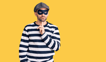 Young handsome man wearing burglar mask thinking concentrated about doubt with finger on chin and...
