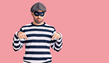 Young handsome man wearing burglar mask pointing down looking sad and upset, indicating direction with fingers, unhappy and depressed.