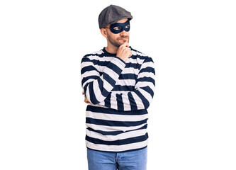 Young handsome man wearing burglar mask with hand on chin thinking about question, pensive expression. smiling with thoughtful face. doubt concept.
