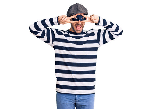 Young handsome man wearing burglar mask doing peace symbol with fingers over face, smiling cheerful showing victory