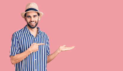 Young handsome man with beard wearing summer hat and striped shirt amazed and smiling to the camera while presenting with hand and pointing with finger.