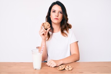 Young beautiful brunette woman drinking a glass of fresh milk with cookies thinking attitude and sober expression looking self confident