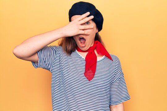 Young beautiful blonde woman wearing french fashion style with scarf and beret peeking in shock covering face and eyes with hand, looking through fingers afraid