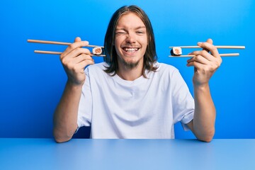 Handsome caucasian man with long hair eating sushi using chopsticks winking looking at the camera...