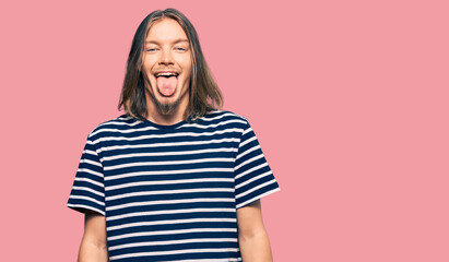 Handsome caucasian man with long hair wearing casual striped t-shirt sticking tongue out happy with funny expression. emotion concept.
