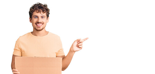 Young handsome man with curly hair holding blank cardboard empty banner smiling happy pointing with hand and finger to the side
