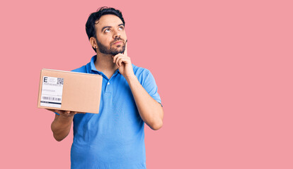 Young hispanic man holding delivery package serious face thinking about question with hand on chin,...