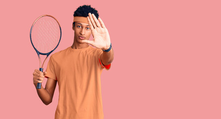Young african american man playing tennis holding racket with open hand doing stop sign with serious and confident expression, defense gesture
