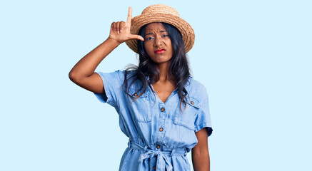 Young indian girl wearing summer hat making fun of people with fingers on forehead doing loser gesture mocking and insulting.