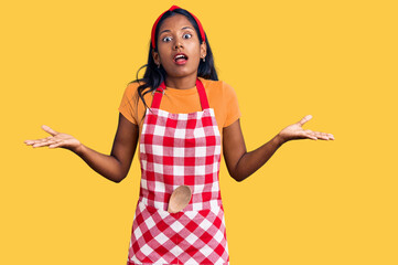 Young indian girl wearing professional baker apron clueless and confused expression with arms and hands raised. doubt concept.