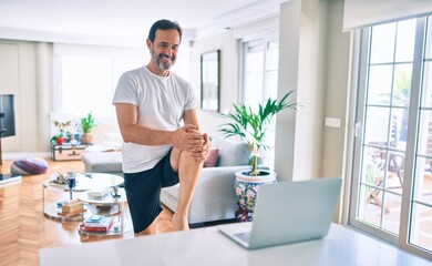 Middle age man with beard training and stretching doing exercise at home looking at sport video on...