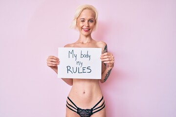 Young blonde woman with tattoo standing shirtless holding banner with my body my rules message winking looking at the camera with sexy expression, cheerful and happy face.