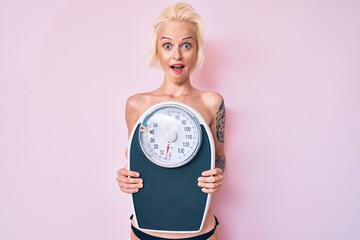 Young blonde woman with tattoo standing shirtless holding weighing machine celebrating crazy and amazed for success with open eyes screaming excited.