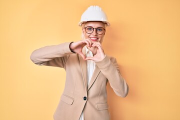 Young blonde woman wearing architect hardhat smiling in love doing heart symbol shape with hands. romantic concept.