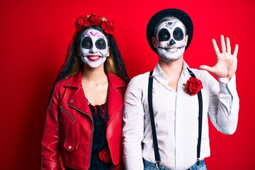 Couple wearing day of the dead costume over red showing and pointing up with fingers number five while smiling confident and happy.