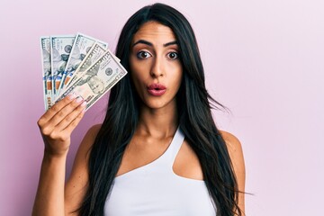 Beautiful hispanic woman holding dollars scared and amazed with open mouth for surprise, disbelief face