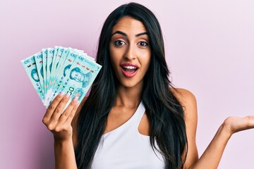 Beautiful hispanic woman holding yuan chinese banknotes celebrating achievement with happy smile and winner expression with raised hand