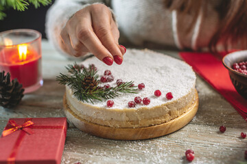 A woman is decorating a white mousse festive cake with red berries sprinkled with coconut flakes that imitating snow. Christmas and New year concept