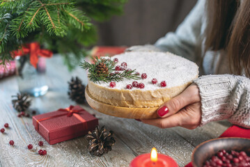 Woman is holding a festive white mousse cake covered with coconut flakes imitating snow and decorated with red berries, cones and fir branches. Concept of the New year and Christmas