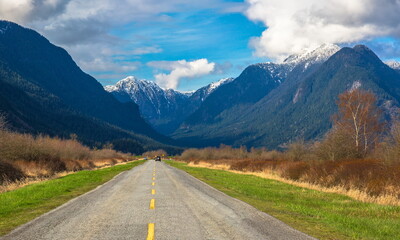 A country road in the Pitt River Valley runs through farm fields and forest to  Pitt Lake on the background of the snow-covered mountain range and a beautiful cloudy sky
