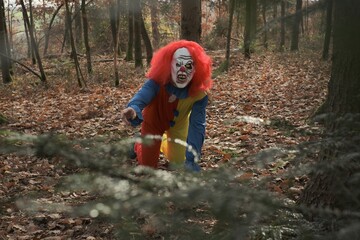 Halloween holiday concept. Horror and fear concept. Scary aggressive clown with red hair in the autumn dark forest. Masquerade and carnival. Scary clown costumes. Creepy clown in the autumn forest
