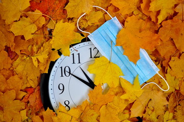 Wall clock and medical mask on the background of yellow autumn leaves.