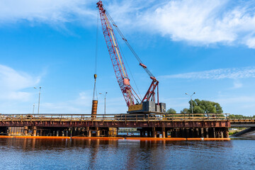 Fototapeta premium Construction of a new bridge. A crane on the background of a blue sky above the lake lifts a metal pipe