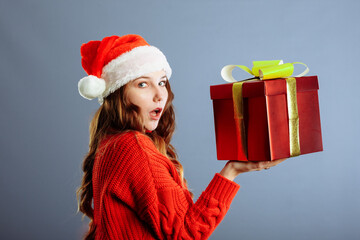 Beautiful Caucasian girl in dress and Santa hat is holding presents, looking at camera and smiling, isolated on gray background